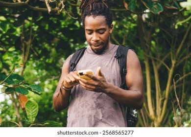 Focused young ethnic man in casual clothes with dreadlocks walking along green tropical forest and browsing web in soft daylight