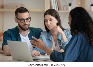 Focused Young Family Couple Listening To African American Realtor At Office. Mixed Race Financial Advisor Real Estate Agent Lawyer Showing Service Presentation On Laptop To Concentrated Spouses.