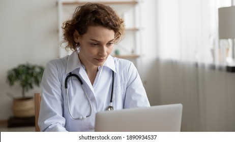 Focused Young Female 35s Doctor Physician In White Coat Working On Computer, Consulting Patients Distantly Online At Clinic Office, Web Surfing Medical Information Or Prescribing Illness Treatment.