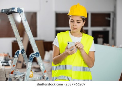 Focused young female foreman wearing yellow vest and safety helmet using smartphone at construction site indoors.. - Powered by Shutterstock