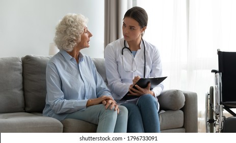 Focused young female general practitioner in white uniform listening to older mature patient complaints, filling medical form at home meeting, doctor examining elderly woman at checkup visit at home. - Powered by Shutterstock