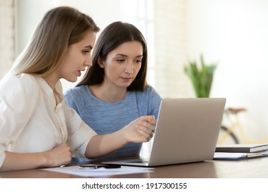 Focused young female leader helping newbie colleague with software applications, working on computer in office. Two serious busy teammates developing online marketing strategy or discussing project.