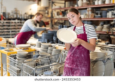 Focused young female potter examining newly crafted ceramic plate, considering subtle nuances of shape and surface in artisanal workshop.. - Powered by Shutterstock