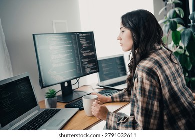 Focused young female software developer working from a home office with multiple monitors and a laptop, reviewing code while holding a coffee mug; productive remote work, technology and programming concept.