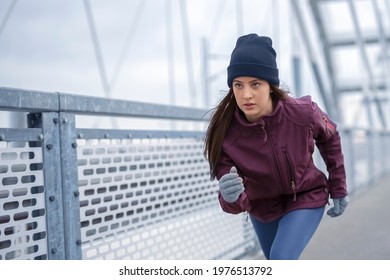 Focused young fitness woman in winter sportswear, winter skully hat and winter gloves jog on bridge during cold day