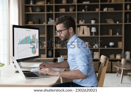 Focused young freelance business man analyzing financial data on desktop large display, working on marketing chart, sales report, using modern Internet technology for job, typing on laptop
