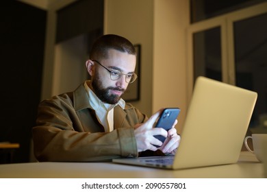 Focused young front end developer or qa engineer testing app on smartphone workloaded, work overtime in empty coworking space before project deadline. Workaholic programmer with smartphone and laptop
