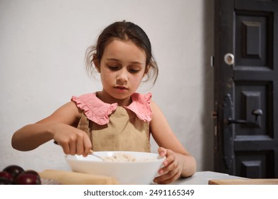 A focused young girl in a pink and beige outfit sits at a table, mixing ingredients in a bowl, learning baking skills in a home kitchen setting. - Powered by Shutterstock