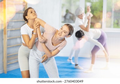 Focused young girl practicing basic protection skills in mock bout with female opponent during self defense course in gym, trying to escape rear grab attack technique, striking chin with elbow