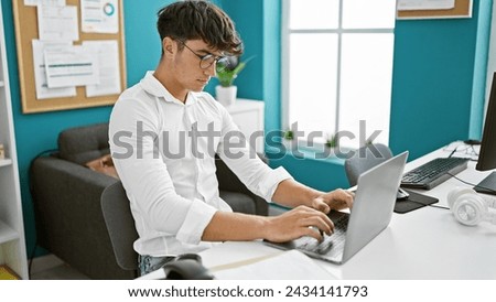 Focused young hispanic teenager, an elegant business worker, using his technology gadget, a laptop, in an indoor office. professional work personified, a portrait of success.