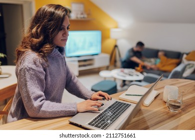 Focused Young Hispanic Woman Sitting At Her Kitchen Table At Home Working On Her Small Business With A Laptop. Mother Working From Home While Her Family Is Playing In The Background.