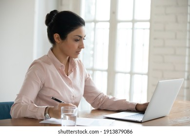 Focused Young Indian Business Woman Working On Computer And Writing Notes. Serious Office Employee Using Laptop At Desk. Female Professional Checking Document. Online Learning, Expertise Concept