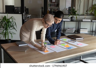 Focused young indian employee managing working processes with smart skilled 30s blonde female boss, using colorful stickers on paper documents at office table, teamwork brainstorming activity.