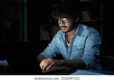 Focused Young Indian Ethnic Business Man Developer Or Web Designer Wearing Glasses Typing On Laptop Distance Working Online Late At Night At Home Or In Dark Office Using Laptop Sitting At Desk.