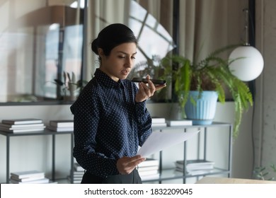 Focused Young Indian Female Employee Read Document Record Voice Message On Smartphone. Millennial Ethnic Businesswoman Hold Paperwork Talk Speak On Speakerphone, Use Digital Assistant On Gadget.