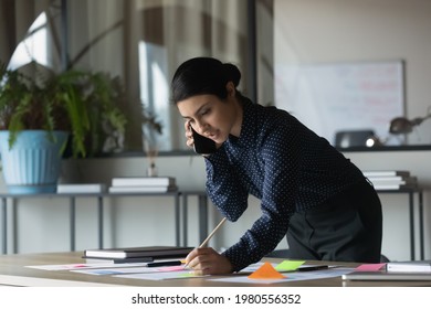 Focused young Indian female employee talk speak on smartphone prepare business paperwork project in office. Serious ethnic businesswoman have cellphone call work on financial startup project. - Powered by Shutterstock