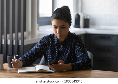 Focused Young Indian Woman Write In Notebook Study Distant At Home Using Smartphone Gadget. Smart Concentrated Ethnic Female Make Notes In Notepad, Look At Cellphone Screen Planning Thinking.