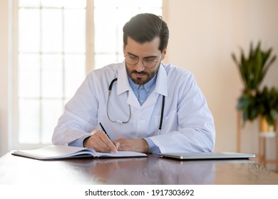 Focused young male doctor in eyewear and medical coat working with paper registry book, making notes, managing patients visits, recording healthcare treatment, sitting at table in clinic office. - Powered by Shutterstock