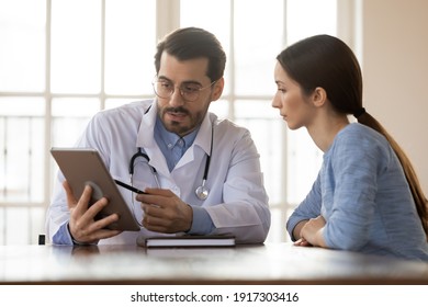 Focused young male doctor pointing at digital computer tablet screen, sharing health tests laboratory results to serious female patient or explaining medical insurance benefits at meeting in clinic.