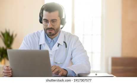Focused young male doctor in wireless headphones with microphone involved in online educational webinar, teaching or learning distantly in clinic office, consulting patient remotely, copy space.