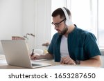 Focused young man businessman company worker employee in glasses wearing wireless headphones, watching educational webinar lecture seminar on laptop online, writing down notes in modern office.