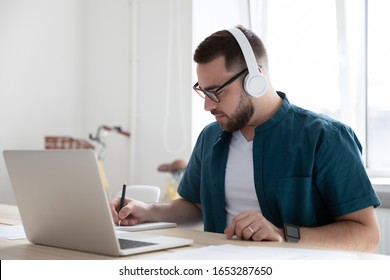 Focused young man businessman company worker employee in glasses wearing wireless headphones, watching educational webinar lecture seminar on laptop online, writing down notes in modern office. - Shutterstock ID 1653287650