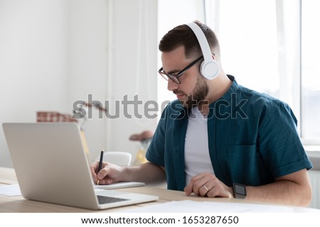Focused young man businessman company worker employee in glasses wearing wireless headphones, watching educational webinar lecture seminar on laptop online, writing down notes in modern office.
