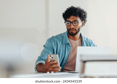 Focused young man in a casual denim shirt engaging with technology in a bright office setting. He is reading a text message from a smartphone, reflecting modern professionalism and productivity.