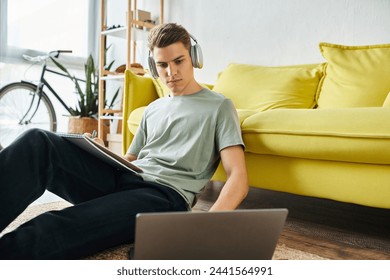 focused young man with headphones on floor near yellow couch studying in laptop and writing in note