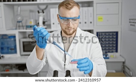 A focused young man in labcoat performs an experiment in a laboratory, pipetting a blue substance into a petri dish.