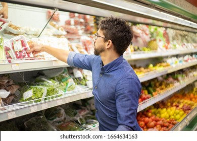 Focused Young Man Shopping In Grocery Store. Side View Of Serious Young Man In Eyeglasses Choosing Fresh Vegetables In Supermarket. Shopping Concept