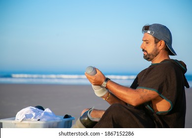 Focused Young Man Sitting On Beach, Holding Ankle Prosthesis. Side View. Artificial Limb And Outdoor Activity Concept