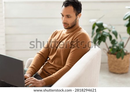 Focused Young Man Using Laptop With Earbuds At Home, Modern Interior