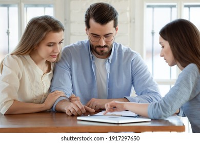 Focused Young Married Couple Discussing Agreement With Skilled Real Estate Agent Or Broker. Professional Financial Advisor Or Saleswoman Explaining Contract Details With Interested Clients At Meeting.