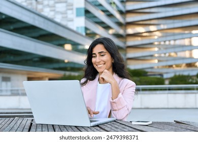 Focused young middle eastern Israel businesswoman using laptop pc online application for work sitting outdoors. Indian or arabic woman manager in business suit doing data base research on computer