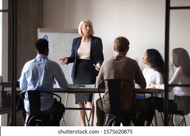 Focused young mixed race colleagues listening to middle aged female boss presenting marketing strategy near whiteboard. Confident older businesswoman speaker giving educational workshop at office. - Shutterstock ID 1575620875