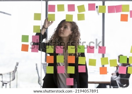 Focused young office employee working on project, using glass board, colorful paper tickers, writing notes. Business coach woman preparing presentation. Scrum management concept