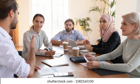 Focused Young And Older Mixed Race Colleagues Gathered At Table, Listening To Male Team Leader Project Explanation. Concentrated Group Of Diverse Business Partners Discussing Development Strategy.