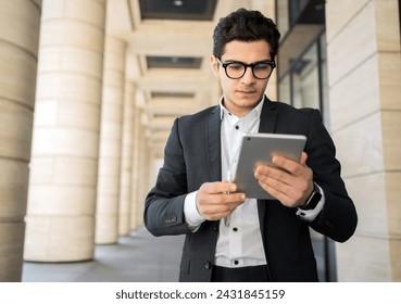 Focused young professional using tablet in front of corporate building, tech-savvy and serious.