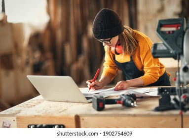 Focused young skilled craftswoman making notes in papers while standing at table with laptop and carpentry instruments in workshop