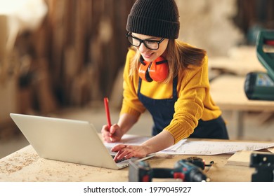 Focused young skilled craftswoman making notes in papers while standing at table with laptop and carpentry instruments in workshop
