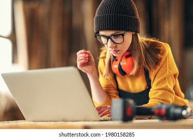 Focused young skilled craftswoman making notes in papers while standing at table with laptop and carpentry instruments in workshop