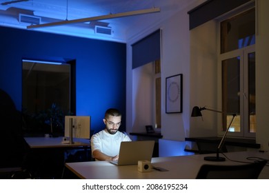 Focused Young Web Designer In Eyeglasses Stay In Office Overtime Work On Project At Dark Night. Freelancer Man, Remote Worker Or Outsourcing Specialist Concentrated On Programming In Laptop Computer