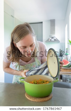 Focused young woman cooking in her kitchen, opening saucepan and smelling vegetable meal. Vertical shot. Cooking at home concept