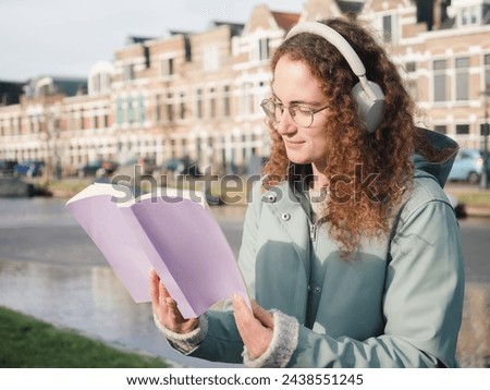A focused young woman with curly hair and glasses, wearing headphones, reads a book by a canal, with classical Dutch architecture in the background