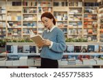 A focused young woman in glasses and a blue sweater reads a book while standing in a vibrant, well-stocked bookstore. The shelves are filled with a variety of books.