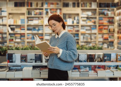 A focused young woman in glasses and a blue sweater reads a book while standing in a vibrant, well-stocked bookstore. The shelves are filled with a variety of books. - Powered by Shutterstock