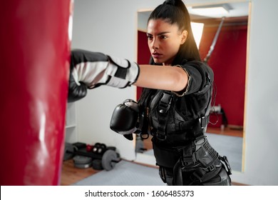 Focused young woman punching boxing bag in ems suit 