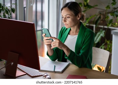 Focused Young Woman Scrolls Social Media Undergoing Online Training And Reads Materials About Business Project. Serious Female Rests From Hard Work, Surfing Internet On Mobile Phone In Cozy Workplace.