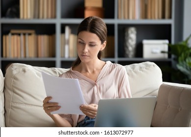 Focused Young Woman Sit On Couch In Living Room Working On Laptop Considering Paperwork Correspondence, Serious Millennial Female Read Paper Contract Or Notice Use Computer At Home